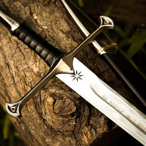 Puede incluir: Espada plateada con empu&ntilde;adura de cuero negro y hoja grabada. La espada presenta un emblema de estrella y descansa sobre una superficie de madera marr&oacute;n texturizada. Tambi&eacute;n se ve una vaina negra.