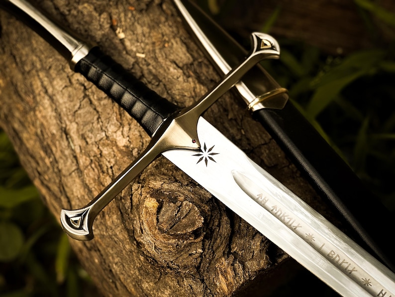 Puede incluir: Espada plateada con empu&ntilde;adura de cuero negro y hoja grabada. La espada presenta un emblema de estrella y descansa sobre una superficie de madera marr&oacute;n texturizada. Tambi&eacute;n se ve una vaina negra.