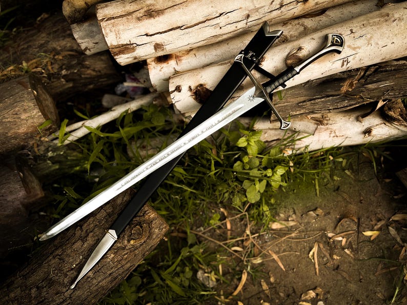 Puede incluir: Dos espadas con hojas plateadas y empu&ntilde;aduras negras descansan sobre una pila de troncos. Una espada est&aacute; envainada en una vaina negra. Las hojas tienen dise&ntilde;os grabados. La escena est&aacute; ambientada al aire libre con follaje verde.