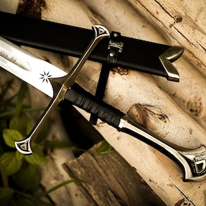 Puede incluir: Imagen detallada de una espada con hoja plateada y detalles dorados. La espada descansa sobre troncos de madera, con una vaina de cuero negro. El mango est&aacute; envuelto en cuero negro y la hoja tiene un dise&ntilde;o de estrella grabado.