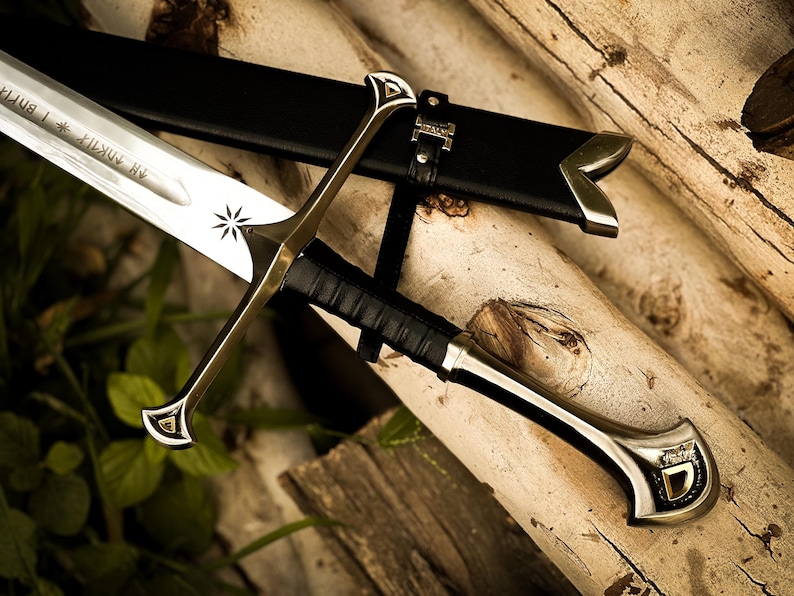 Puede incluir: Imagen detallada de una espada con hoja plateada y detalles dorados. La espada descansa sobre troncos de madera, con una vaina de cuero negro. El mango est&aacute; envuelto en cuero negro y la hoja tiene un dise&ntilde;o de estrella grabado.