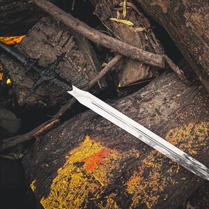 K&ouml;nnte beinhalten: Ein Schwert mit schwarzem Griff und kunstvoller Parierstange liegt auf einem Stapel verwitterter Baumst&auml;mme. Die silberne Klinge reflektiert das Licht und bildet einen Kontrast zum dunklen Griff und der Holzmaserung.