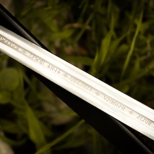 Puede incluir: Primer plano de una hoja de espada plateada con texto grabado. La hoja est&aacute; en &aacute;ngulo, reflejando la luz, y sobre un fondo borroso verde y negro. La espada parece ser un art&iacute;culo hist&oacute;rico o de fantas&iacute;a.
