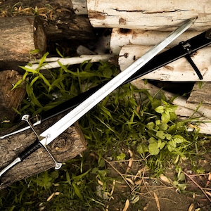 Puede incluir: Una espada plateada con empu&ntilde;adura negra y vaina negra descansa sobre una pila de troncos y follaje verde. La espada tiene una hoja de doble filo y una guarda decorativa. La vaina es de cuero.