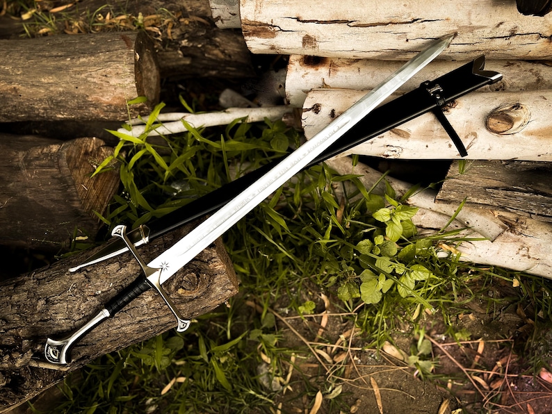 Puede incluir: Una espada plateada con empu&ntilde;adura negra y vaina negra descansa sobre una pila de troncos y follaje verde. La espada tiene una hoja de doble filo y una guarda decorativa. La vaina es de cuero.