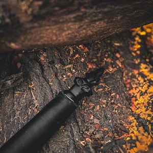 K&ouml;nnte beinhalten: Ein schwarzer Flaschenverschluss mit dekorativer Spitze, vor einem strukturierten, dunkelbraunen Hintergrund. Der Verschluss hat eine verzierte, spitze Spitze. Das Bild hat einen warmen, erdigen Ton.
