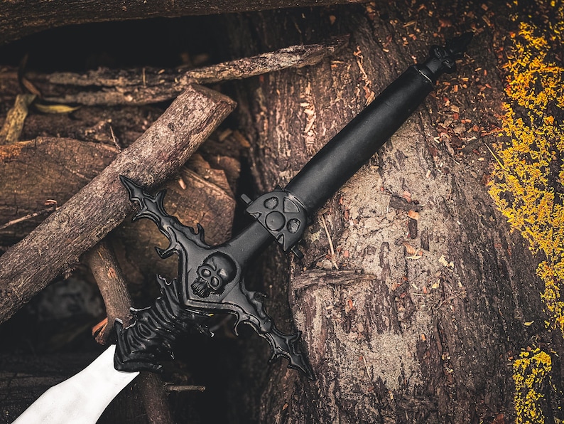 K&ouml;nnte beinhalten: Ein schwarzes Fantasy-Schwert mit einem Totenkopf-Design am Knauf und einer wei&szlig;en Klinge. Das Schwert ist vor einem Hintergrund aus Holzst&auml;mmen und gelbem Laub platziert, was einen dramatischen Kontrast erzeugt.
