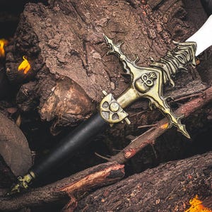 K&ouml;nnte beinhalten: Ein dekoratives Schwert mit schwarzem Griff und goldfarbener Parierstange mit Totenkopf-Design. Die Klinge ist wei&szlig;. Das Schwert liegt auf einem Stapel Holzscheite und erzeugt eine rustikale &Auml;sthetik.