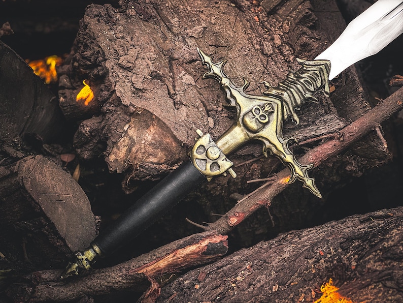 K&ouml;nnte beinhalten: Ein dekoratives Schwert mit schwarzem Griff und goldfarbener Parierstange mit Totenkopf-Design. Die Klinge ist wei&szlig;. Das Schwert liegt auf einem Stapel Holzscheite und erzeugt eine rustikale &Auml;sthetik.
