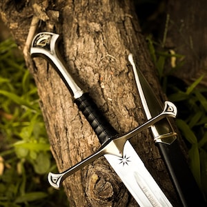 Puede incluir: Espada de fantas&iacute;a con una hoja plateada y un mango de cuero negro, apoyada en un tronco de &aacute;rbol. La espada presenta un emblema de estrella y va acompa&ntilde;ada de su vaina negra. El dise&ntilde;o evoca una est&eacute;tica medieval o fant&aacute;stica.
