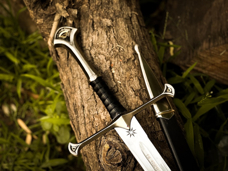 Puede incluir: Espada de fantas&iacute;a con una hoja plateada y un mango de cuero negro, apoyada en un tronco de &aacute;rbol. La espada presenta un emblema de estrella y va acompa&ntilde;ada de su vaina negra. El dise&ntilde;o evoca una est&eacute;tica medieval o fant&aacute;stica.