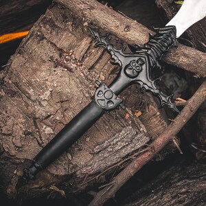 K&ouml;nnte beinhalten: Ein schwarzes Fantasy-Schwert mit einem verzierten Knauf mit Totenkopf-Design und einer wei&szlig;en Klinge. Das Schwert liegt auf einem Haufen verwitterter Holzscheite und &Auml;ste und erzeugt eine rustikale und dramatische Szene.
