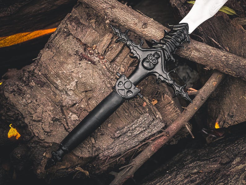 K&ouml;nnte beinhalten: Ein schwarzes Fantasy-Schwert mit einem verzierten Knauf mit Totenkopf-Design und einer wei&szlig;en Klinge. Das Schwert liegt auf einem Haufen verwitterter Holzscheite und &Auml;ste und erzeugt eine rustikale und dramatische Szene.