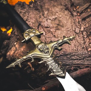 K&ouml;nnte beinhalten: Ein dekoratives Schwert mit schwarzem Griff und goldfarbener Parierstange mit Totenkopf-Design. Die Klinge ist silberfarben. Das Schwert liegt auf einer Holzoberfl&auml;che, was auf ein Fantasy- oder historisches Thema hindeutet.