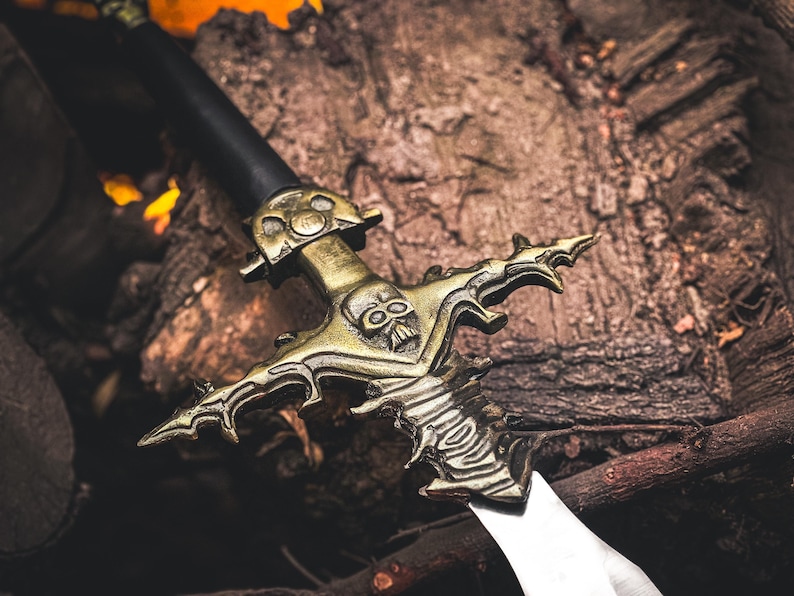 K&ouml;nnte beinhalten: Ein dekoratives Schwert mit schwarzem Griff und goldfarbener Parierstange mit Totenkopf-Design. Die Klinge ist silberfarben. Das Schwert liegt auf einer Holzoberfl&auml;che, was auf ein Fantasy- oder historisches Thema hindeutet.