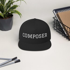 Può includere: Cappellino da baseball nero con visiera piatta, con la scritta "COMPOSER" in lettere bianche sul davanti. Il cappello è appoggiato su una superficie bianca. Sullo sfondo, una pianta in vaso e un cestino di filo metallico.