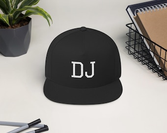 以下が含まれることがあります： 前面に白い文字「DJ」が刺繍された黒いフラットブリムの野球帽。 白い表面に置かれ、鉢植えの植物、鉛筆、ワイヤーバスケットが背景にあります。