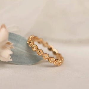 Puede incluir: Un anillo dorado con un diseño floral de margaritas repetidas. El anillo se encuentra sobre un fondo suave y borroso, resaltando el delicado patrón floral. El diseño del anillo es una banda continua de pequeñas flores detalladas.