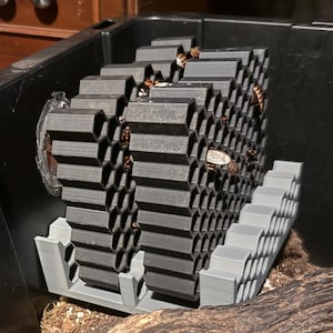 May include: A gray, 3D-printed insect habitat with a honeycomb design. The habitat is inside a black plastic container, with a wooden log and cardboard pieces visible. The habitat is designed to house insects.