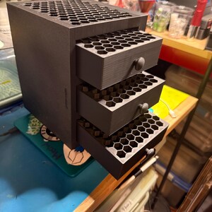 May include: A matte black storage unit featuring three pull-out drawers, each with a honeycomb-patterned interior grid. The exterior of the unit and the top surface also display a matching hexagonal honeycomb design, with small round knobs on each drawer.