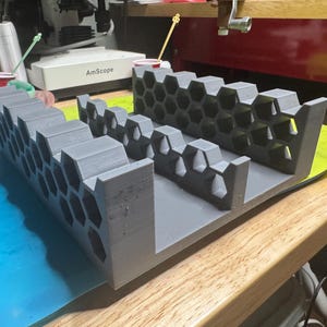 Peut inclure: Un organisateur gris imprimé en 3D avec un motif en nid d'abeille. L'organisateur comporte plusieurs compartiments et est conçu pour contenir de petits objets. L'article est posé sur une surface bleue et jaune.