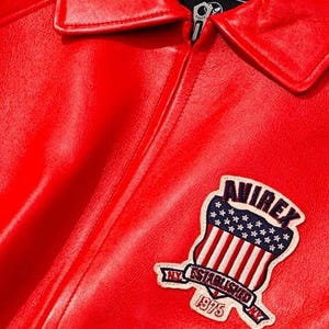 May include: Close-up of a bright red leather jacket with a classic collar and a silver zipper. The jacket features a patch with the text "AVIREX" above a design of the American flag and the text "NY ESTABLISHED 1975".
