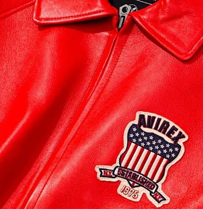 May include: Close-up of a bright red leather jacket with a classic collar and a silver zipper. The jacket features a patch with the text "AVIREX" above a design of the American flag and the text "NY ESTABLISHED 1975".