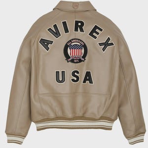 May include: Tan leather jacket with the word "AVIREX" arched across the back in black letters. Below the word "AVIREX" is a circular patch with the words "ALL AMERICANS ESTABLISHED 1975" and a US flag design. The word "USA" is below the patch.