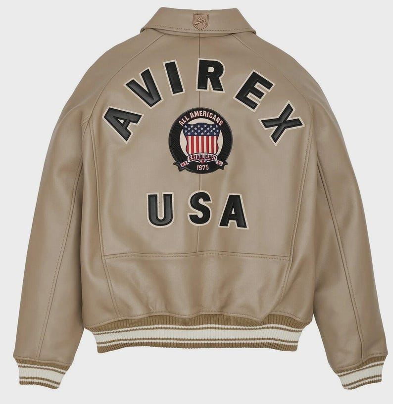 May include: Tan leather jacket with the word "AVIREX" arched across the back in black letters. Below the word "AVIREX" is a circular patch with the words "ALL AMERICANS ESTABLISHED 1975" and a US flag design. The word "USA" is below the patch.