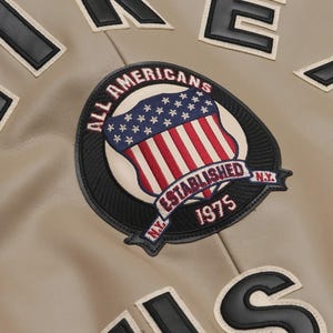 May include: Close-up of a tan varsity jacket with black and white lettering and patches. The jacket features a circular patch with the text "ALL AMERICANS ESTABLISHED 1975 N.Y." and a shield design with the American flag.