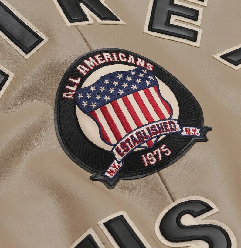 May include: Close-up of a tan varsity jacket with black and white lettering and patches. The jacket features a circular patch with the text "ALL AMERICANS ESTABLISHED 1975 N.Y." and a shield design with the American flag.