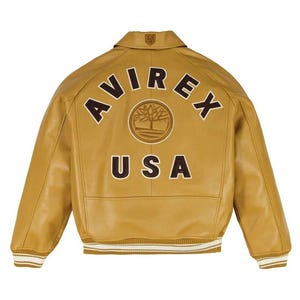 Könnte beinhalten: Senfgelbe Lederjacke mit dem Wort "AVIREX" in braunen Buchstaben auf dem Rücken, über einem kreisförmigen Emblem und dem Wort "USA". Die Jacke hat einen Kragen und gerippte Bündchen und Saum mit weißen Streifen.