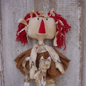 Könnte beinhalten: Eine handgemachte Stoffpuppe mit roten Wollhaaren, einem braunen Kleid und einer weißen Katze in ihren Armen. Die Puppe hat ein bemaltes Gesicht mit einer roten Nase und der Name "Annie" ist auf ihrem Kleid geschrieben.