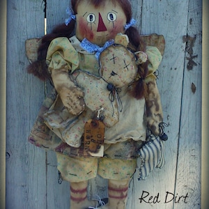 Puede incluir: Una muñeca de trapo hecha a mano con cabello castaño, vestida con un vestido amarillo con estampado floral, sosteniendo un pequeño animal de peluche. La muñeca está de pie frente a un fondo de madera azul. El texto "Old & Worn" es visible en una etiqueta adjunta al vestido de la muñeca. El texto "Red Dirt Primitives" es visible en la esquina inferior derecha de la imagen.