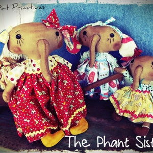 May include: Three handmade fabric dolls with elephant-like features and long noses. The dolls are wearing colorful dresses with floral patterns and are posed on a wooden surface. The text "The Phant Sisters" is written below the dolls.