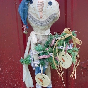 May include: A whimsical, handmade fabric doll with a stitched face, button eyes, and a blue cap. The doll wears a scarf and holds a small wreath with snowman ornaments. The doll is decorated with red and white ribbons and a bell.