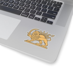 May include: A sticker featuring a golden lion holding a sword, with a sunburst design behind it. The sticker is placed on a silver laptop, with the keyboard visible. The lion is a symbol of strength and heritage.
