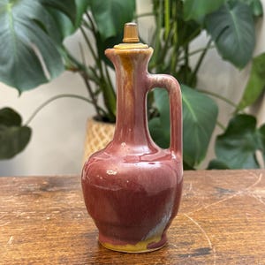 May include: A maroon ceramic oil cruet with a handle and a spout. The cruet has a golden-colored stopper and a yellow base. The cruet is on a wooden surface with green plants in the background. The cruet is approximately 8 inches tall.