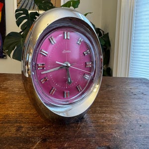 May include: A vintage chrome alarm clock with a vibrant pink face. The clock has a rounded, oval shape and features silver-toned hour markers and hands. The clock face has the brand name "Linden" printed on it. The clock is sitting on a wooden surface.
