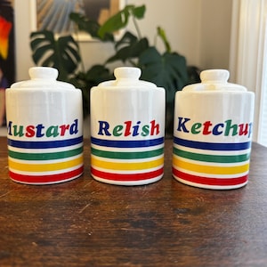 May include: Three white ceramic condiment jars with lids. Each jar has a different word in colorful letters: "Mustard", "Relish", and "Ketchup". Each jar has horizontal stripes in red, yellow, green, blue, and a dark blue stripe.