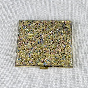 May include: A square compact mirror case featuring a gold-toned metallic frame and a top surface covered in a dense, textured layer of gold glitter mixed with small, multi-colored reflective particles in shades of blue, pink, and green.