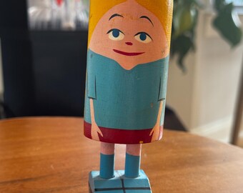 May include: A vintage wooden doll with a painted face, yellow hair, and a blue top. The doll has a red skirt, pink legs, and blue boots. It stands on a blue rectangular base. The doll is a charming example of folk art.