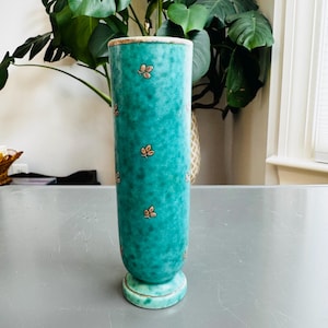 May include: A tall, cylindrical turquoise vase with a textured surface and a flared base. The vase is adorned with small, orange-red floral motifs. The rim and base have a gold-colored trim. The vase is approximately 10 inches tall.