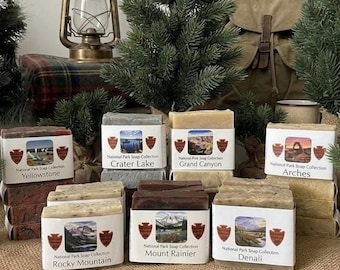 Könnte beinhalten: Eine Sammlung rechteckiger Seifenstücke, jedes in ein weißes Papieretikett mit einem Nationalpark-Bild und dem Text National Park Soap Collection gewickelt. Die Etiketten benennen Parks wie Yellowstone, Crater Lake, Grand Canyon, Arches, Rocky Mountain, Mount Rainier und Denali.