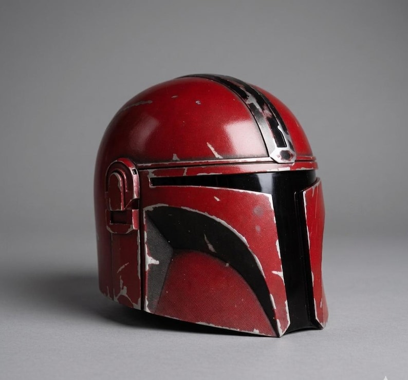 Puede incluir: Un casco mandaloriano rojo y negro con aspecto desgastado. El casco presenta una visera negra y una franja plateada en la parte superior. El casco es un accesorio de la franquicia Star Wars.