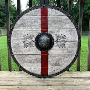 May include: A circular wooden shield featuring a weathered white finish with a vertical red stripe running through the centre. A dark metal rim with rivets surrounds the edge, and a central dark metal boss is flanked by black animal motifs.