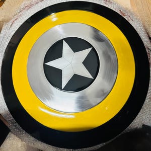 Puede incluir: Un escudo redondo con un anillo exterior negro, un anillo central amarillo y un c&iacute;rculo interior plateado. Una estrella blanca est&aacute; centrada sobre un fondo negro dentro del c&iacute;rculo plateado. R&eacute;plica de escudo de superh&eacute;roe.