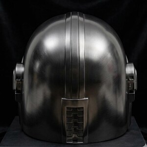 May include: A metallic silver helmet with a rounded top and vertical ridges. The helmet has ear pieces on either side and a rectangular detail at the base. The surface has a brushed metal texture.