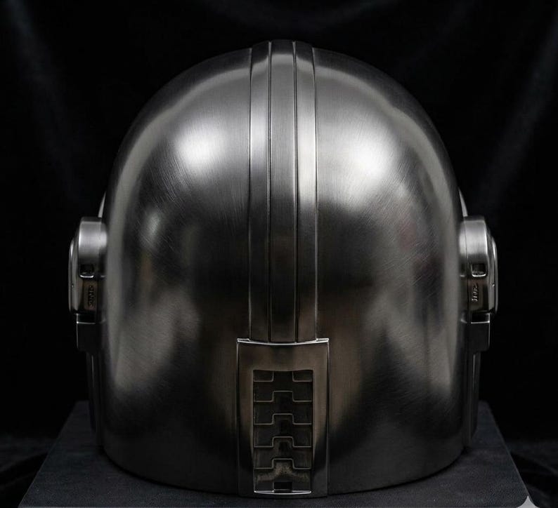 May include: A metallic silver helmet with a rounded top and vertical ridges. The helmet has ear pieces on either side and a rectangular detail at the base. The surface has a brushed metal texture.