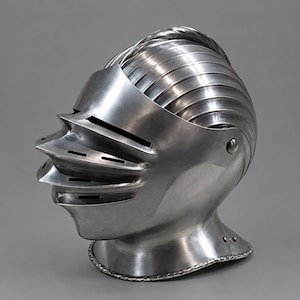 May include: A polished steel close helmet featuring a rounded skull with a prominent central comb and a multi-layered visor. The visor includes three horizontal ventilation slits and a curved, pointed profile extending from the front face.